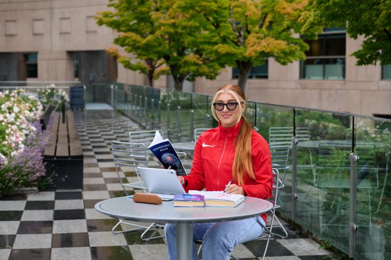 Ashlyn sits at a table outside of a school building with a laptop, books, and notebooks. She is wearing a red jacket and sunglasses, and smiles looking at the camera.