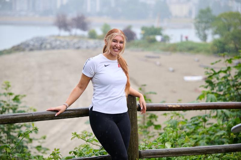 Ashlyn stands outdoors, posing against a log fence. She is smiling at the camera and resting her left arm on top of the fence. She is wearing a white athletic top and black pants