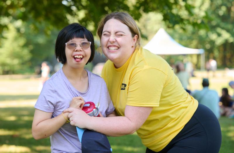 A CNIB child participant with glasses and a CNIB staff member laugh as their photo is taken at an outdoor event.