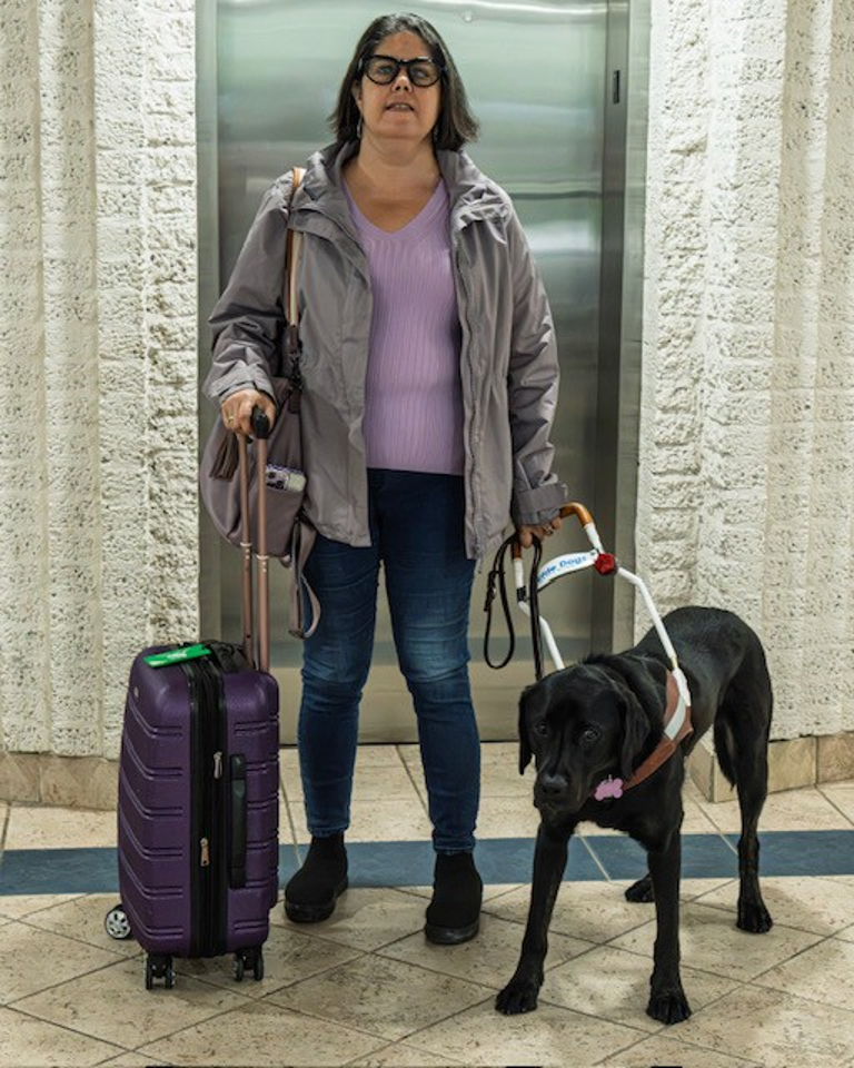 Karoline stands next to her black Labrador guide dog, Guardian, in front of an elevator. Guardian is in harness; Karoline holds the harness handle in her left hand and the handle of a suitcase in her right hand.