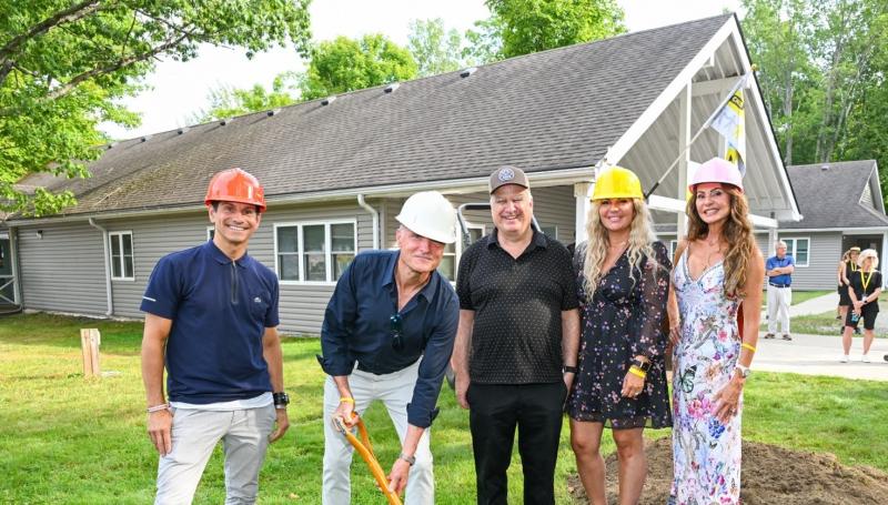 \Five people pose at a groundbreaking ceremony on the lawn in front of a grey building with a peaked roof at CNIB Lake Joe. Three of them wear construction helmets—orange, white, and yellow—and one man with the white helmet is holding a shovel in the ground. The group is smiling, and a few people stand in the background near the building.