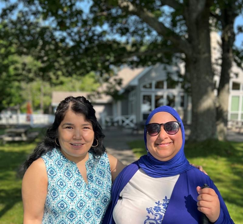 Najla [left] and Salma [right] smile outdoors in front of a leafy tree and a light grey lodge-style building at CNIB Lake Joe.