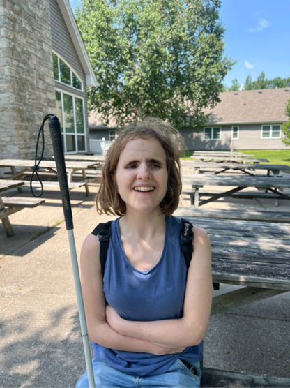 Colombe sits on a wooden picnic bench at CNIB Lake Joe, smiling with her arms crossed. A white cane is propped up beside her. Behind her are several empty picnic tables, a large tree, and a building with stone siding and large windows under a bright blue sky.