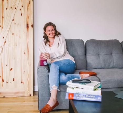 A young woman sitting on a gray couch wearing jeans and a cream coloured sweater. There are books about health stacked on a coffee table in front of her. She is smiling, holding a glass jar filled with fruit juice.
