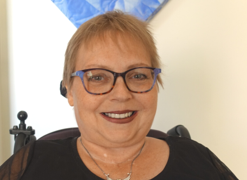 A portrait of Lena smiling at the camera. She has short light-blond hair, blue-framed glasses, a black top, and a silver necklace, and is seated indoors. 