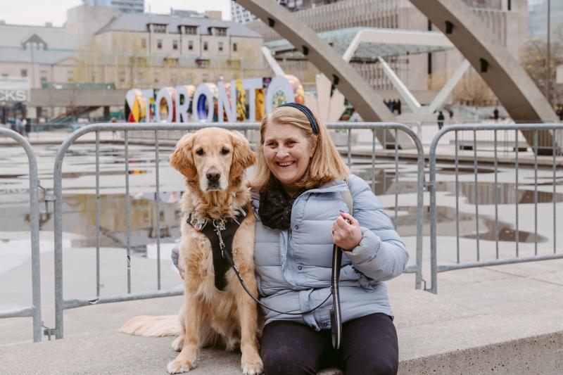 Lindsay sits beside her golden retriever guide dog, Charles, at Nathan Phillips Square in Toronto. Charles is in harness, and Lindsay smiles at the camera with one arm around him. 