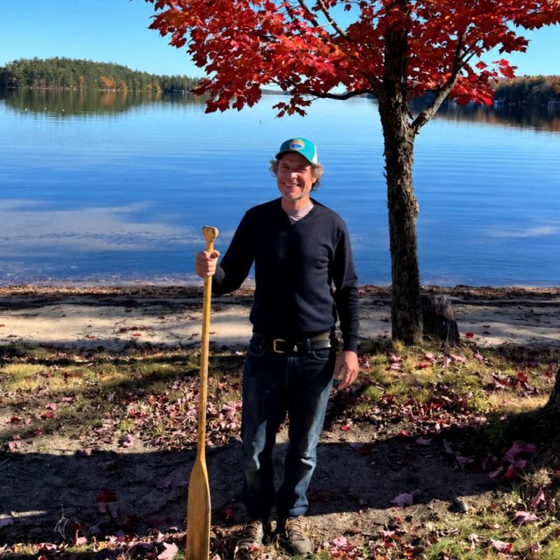 Mark is standing at the shore of a lake smiling at the camera holding a padel in his right hand.