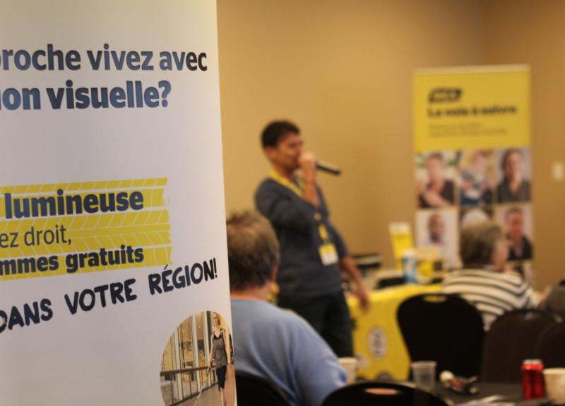 A person holds a microphone and speaks to an audience seated at a CNIB Mobile Hub. Branded informational banners are visible in the background.