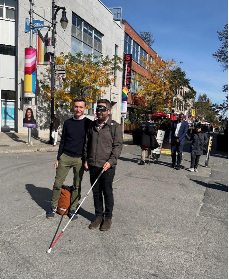 Chris McCray, alongside Robert Beaudry, blindfolded and holding a white cane, stands in the middle of an intersection on Ste-Catherine Street East. Behind them, Shalom Foly and Jean-François Kacou guide and talk with Najla Noori.