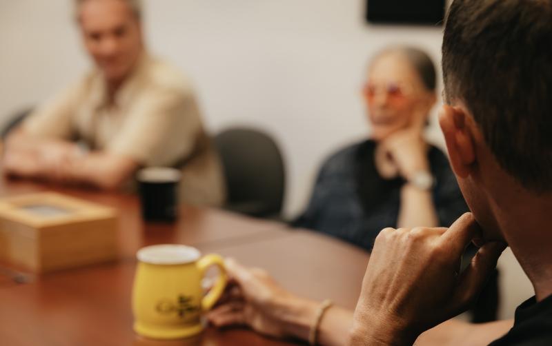 Three people sit around a conference table engaged in discussion. The focus is on a person in the foreground holding a yellow coffee mug. Two other individuals are visible in the background, slightly out of focus, one wearing red-tinted glasses and the other with arms crossed.