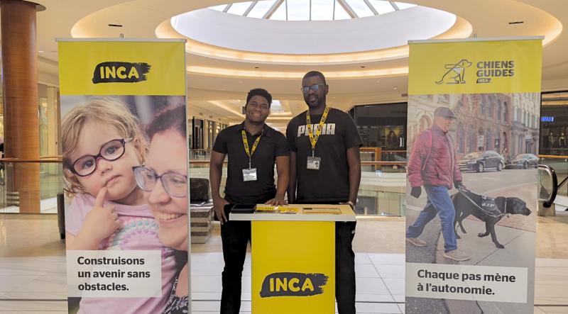 Two canvassers wearing CNIB lanyards stand by a CNIB booth next to CNIB and CNIB Guide Dogs banners inside a mall.