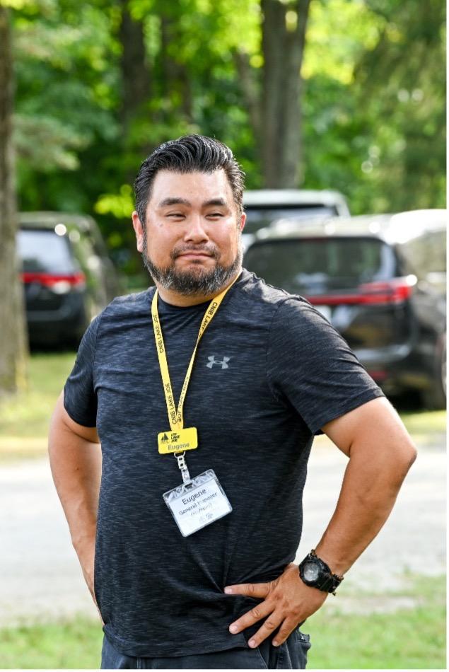 Eugene Chong stands outdoors with his hands on his hips and a slight smile. He has short, swept-back dark hair and a neatly trimmed salt-and-pepper beard. He wears a dark athletic T-shirt, a yellow CNIB Lake Joe lanyard with name badges, and a black wristwatch. Sunlit green trees and parked vehicles are softly blurred behind him.