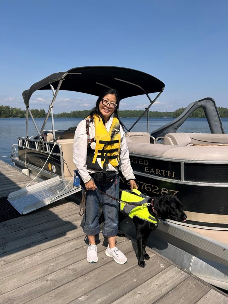 Kathryn is standing on the dock with a pontoon boat behind her. She is wearing a lifejacket and holding her guide dog on harness. It's a bright sunny day and she is smiling at the camera.