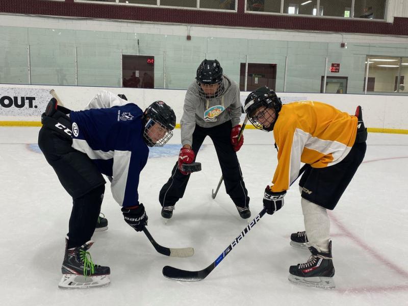 Two blind hockey players hold their sticks against the ice, waiting for the referee who stands between them to drop the puck.