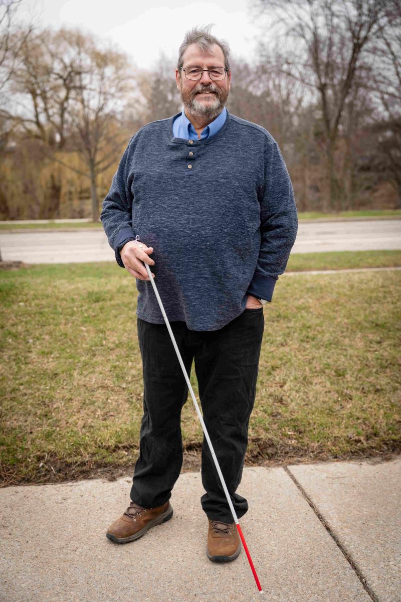 A middle-aged man with grey hair, glasses, and a beard stands smiling on a sidewalk outdoors. He is wearing a navy blue henley sweater over a light blue collared shirt and black pants with brown boots. He holds a white mobility cane with a red tip in his right hand.