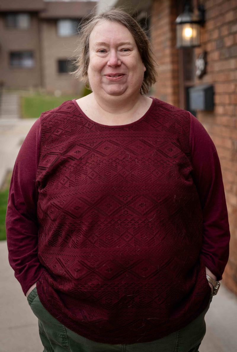 ​​A middle-aged woman with light brown hair stands smiling outdoors on a sidewalk in front of a brick building with a wall-mounted lantern. She is wearing a dark burgundy long-sleeve top with a textured pattern and olive green pants.