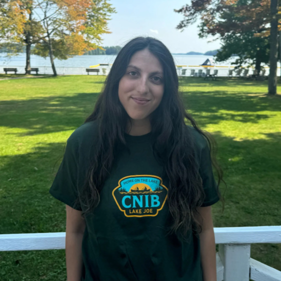 Stela is standing on the CNIB Lake Joe boardwalk with her back to the lake wearing a CNIB Lake Joe olive green t-shirt.