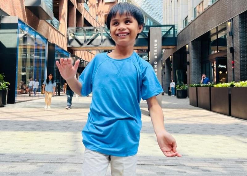 A smiling child in a blue shirt and light shorts stands in an outdoor shopping area, waving.