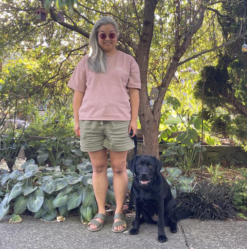 Theresa, wearing a pink shirt and light green shorts, is standing beside a black Labrador retriever, Summer, who is sitting. Theresa and Summer are on a path in front of a garden with leafy green plants.