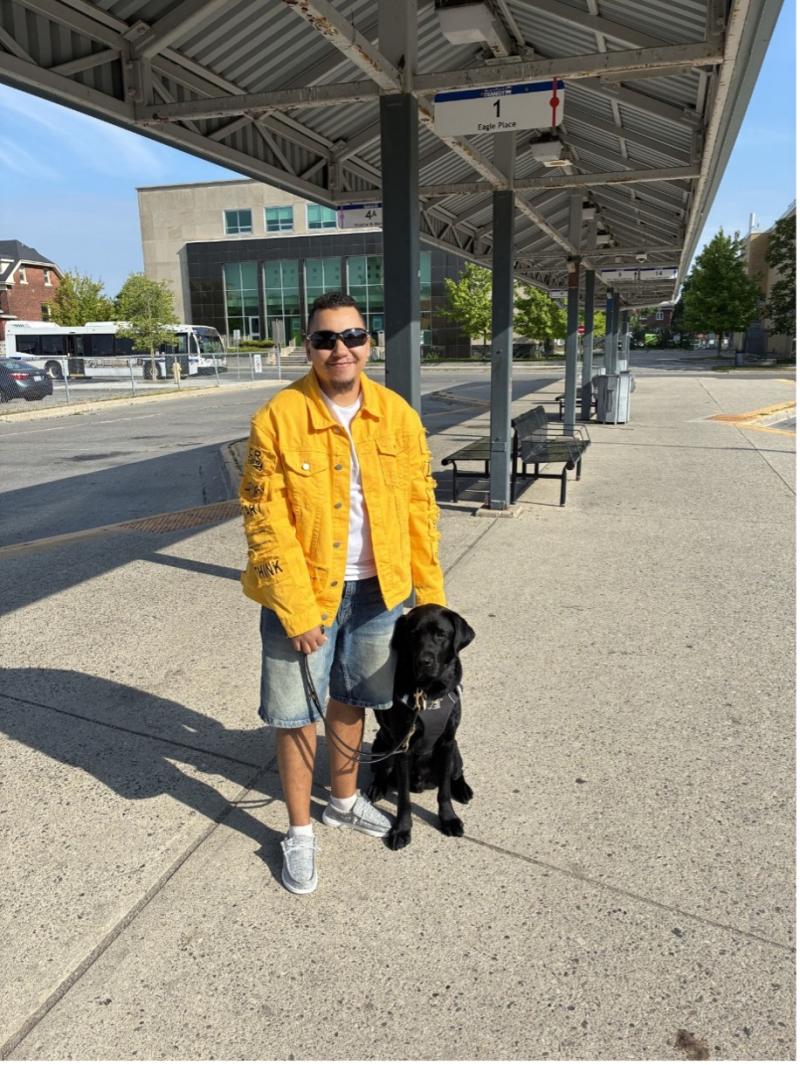 Tye stands beside his black Labrador guide dog, Zed, at an outdoor transit station. Tye hold’s Zed’s harness and leash.&nbsp;
