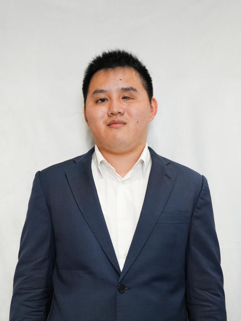 Zhicheng, a young Asian man wearing a navy suit with a white dress shirt, stands in-front of a white background. Zhicheng is smiling and the photograph resembles a professional headshot.