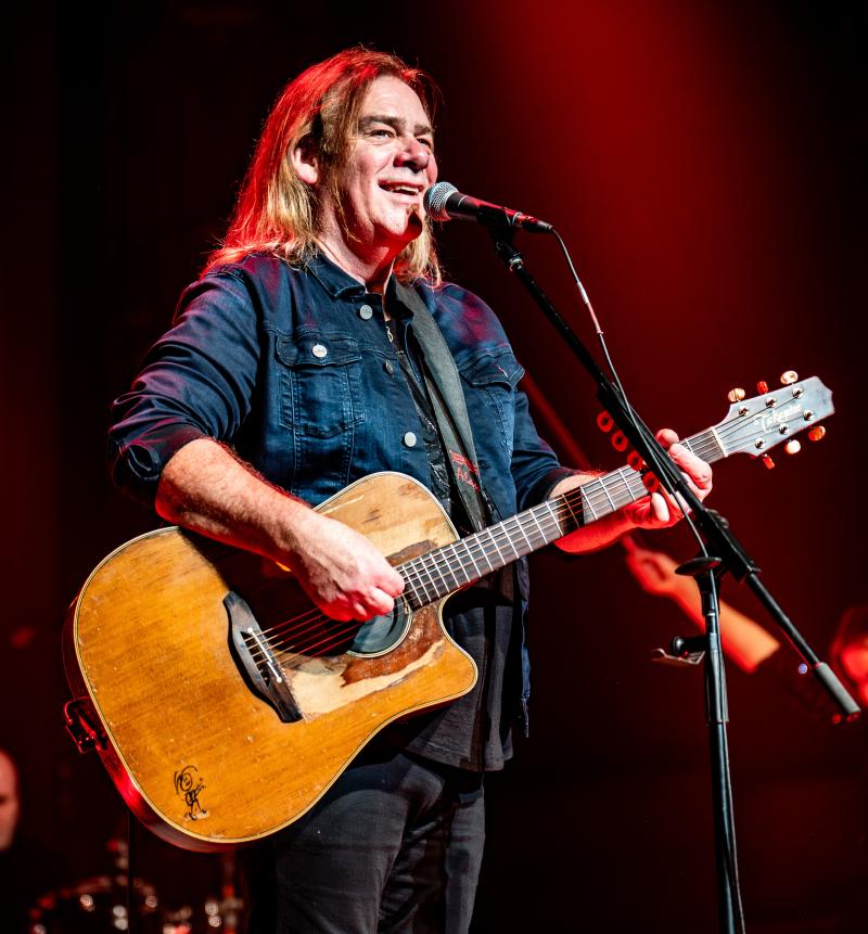 Musician Alan Doyle stands on stage in front of a microphone holding a guitar 