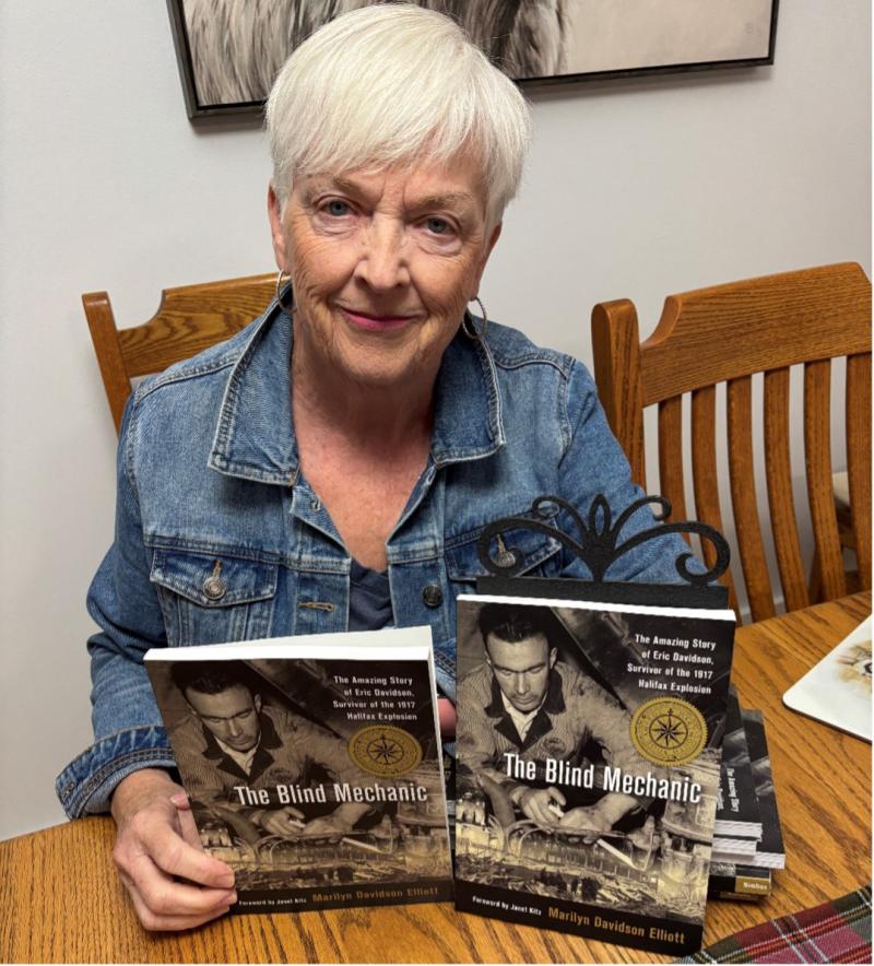 Author Marilyn is sitting at a dining room table holding up copies of her book, “The Blind Mechanic.”