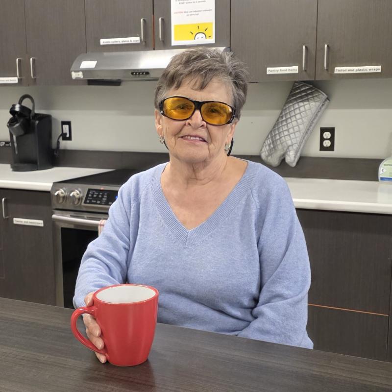 An older woman wearing amber-tinted glasses sits at a table holding a red mug.