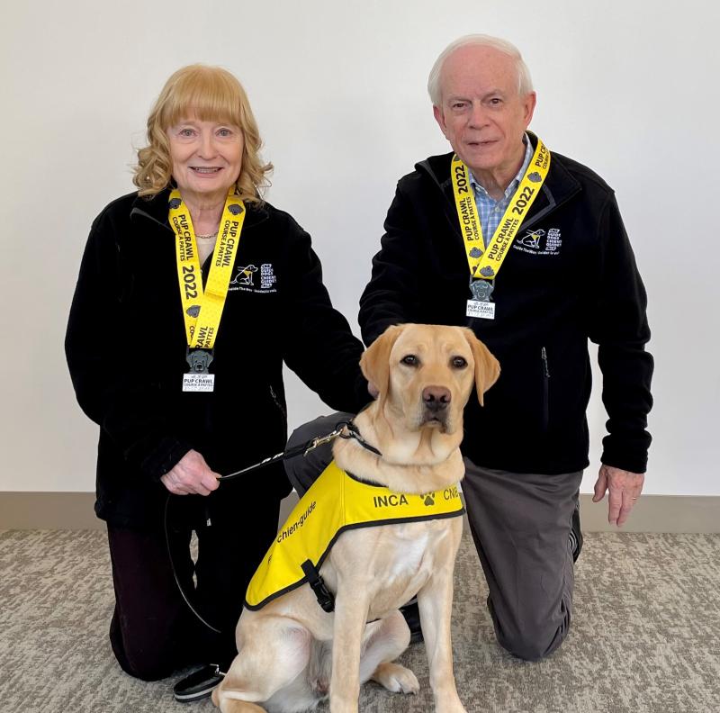 Mary and John Crocker, both smiling, kneel on the ground next to a blonde dog wearing a CNIB "Future Guide Dog" vest