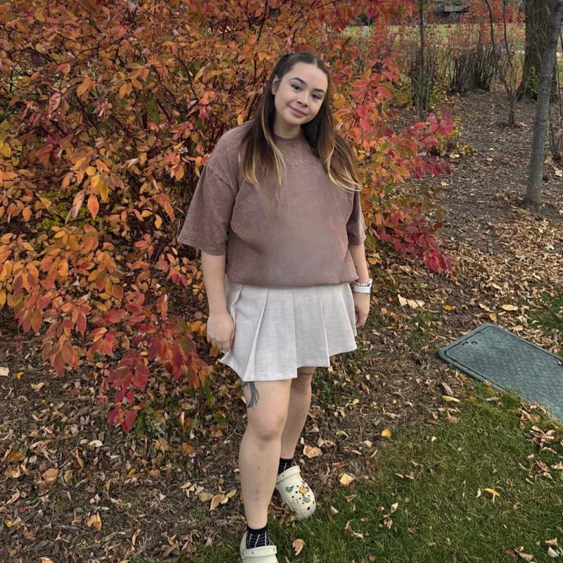 Alexis is standing in front of an autumnal bush wearing a brown t-shirt and beige skirt. She has long brown hair that fades into blond and she is standing with her head on a tilt smiling.