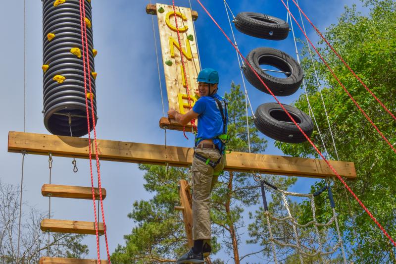 A person wearing a helmet and climbing harnesses scales up the vertical playground at CNIB Lake Joe, which includes wooden beams, rock climbing holds, a rope ladder, and suspended tires.