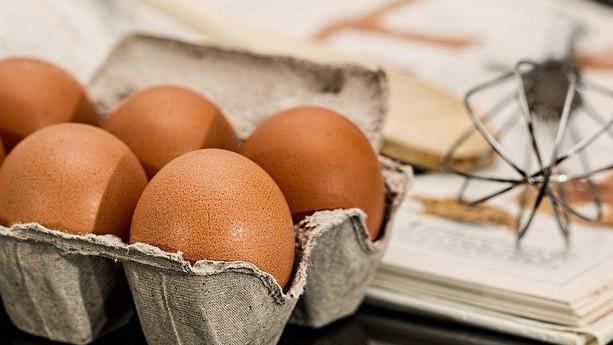 A carton of eggs. A whisk sits on a recipe book