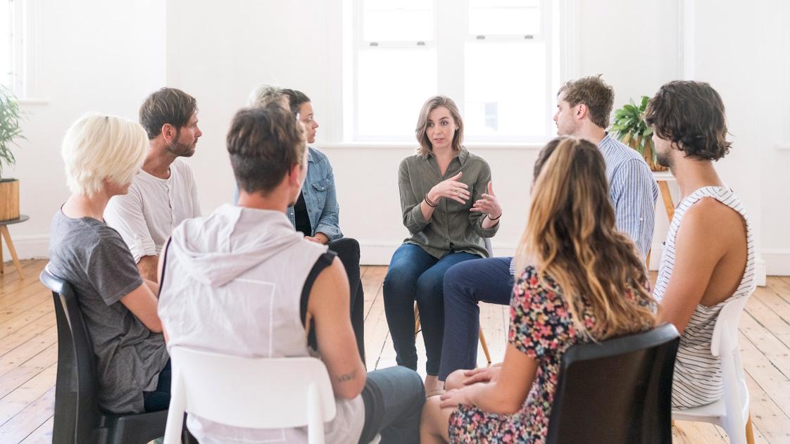 A support group of eight people sit in chairs in a circle and engage in lively conversation. 