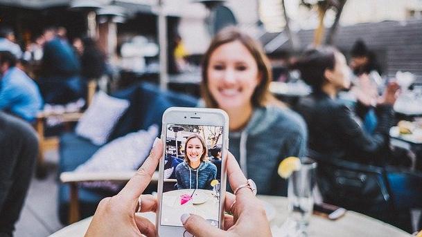 Two women dining. One woman takes a picture of her friend with her smartphone.