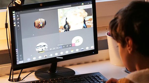 A young child sits at a desktop computer. The computer screen displays a Zoom call.