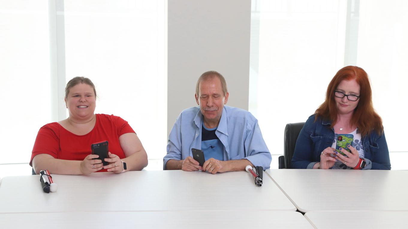 Three people sit with iPhones in hand and white canes sitting on the table in front of them.