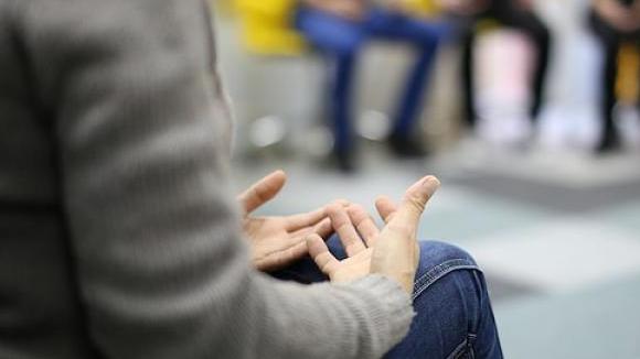 A person sits in a group circle. Their hands are clasped in their lap.
