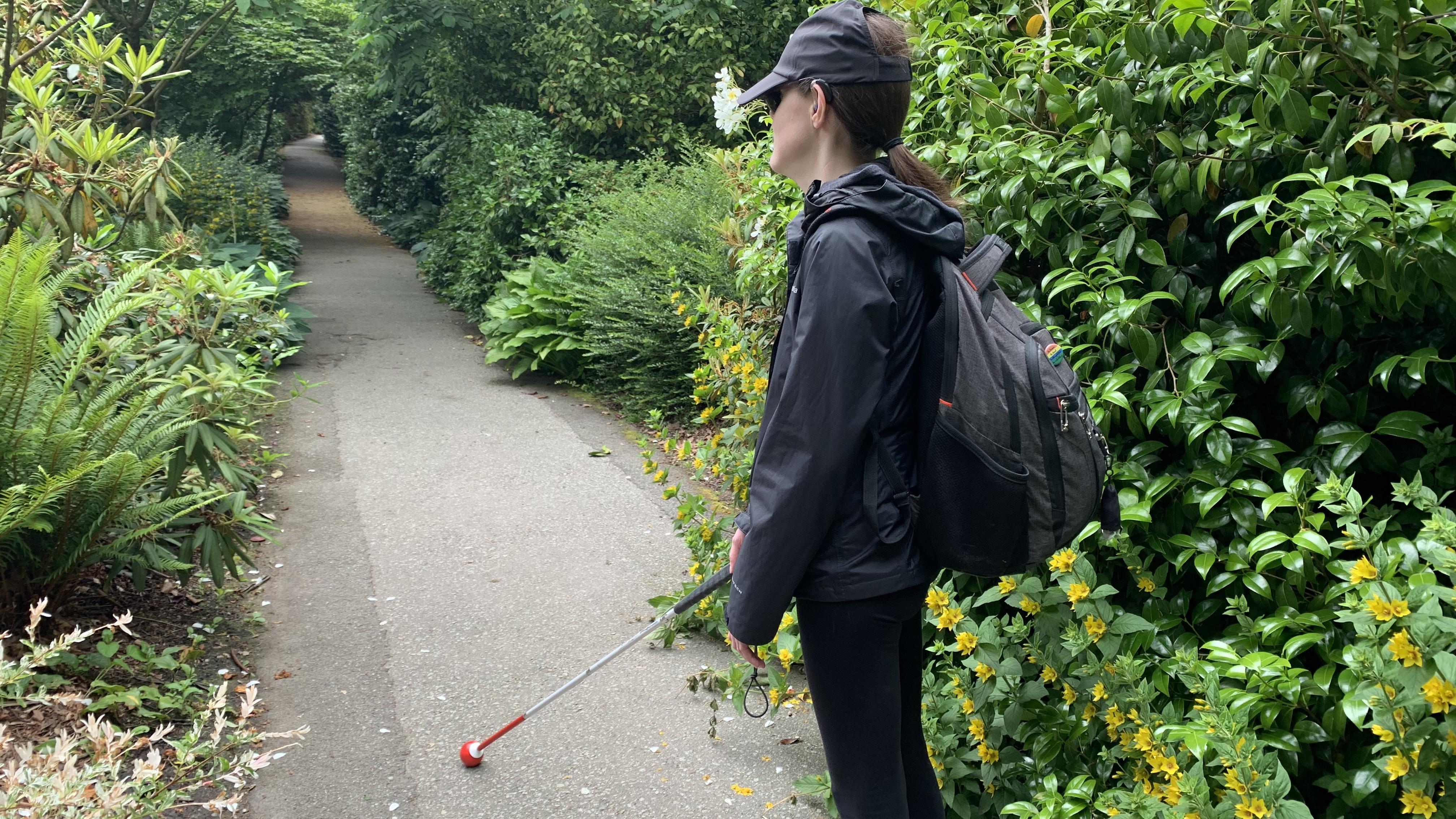 Abby walks along a lush green nature pathway and navigates with their white cane. Abby is turned away from the camera