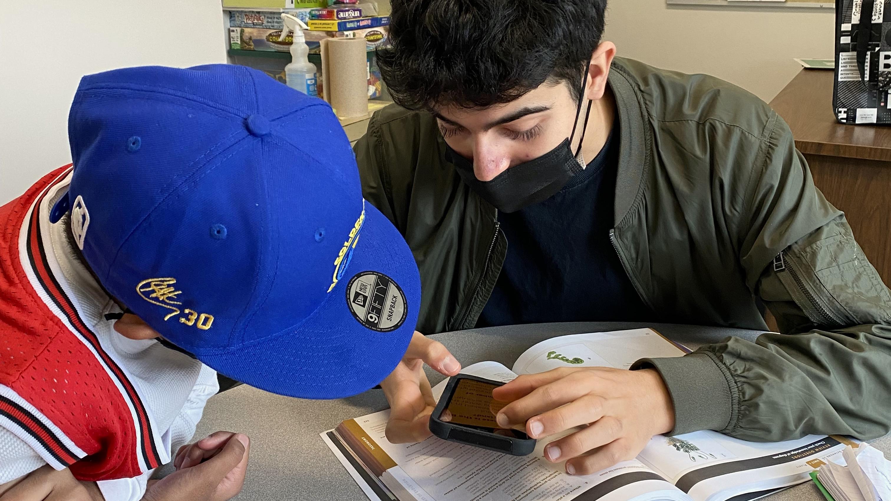 In a classroom, Aidan sits with a young student and shows him the magnification features on his iPhone. Aidan is holding his iPhone over a textbook.  The student intently leans-in for the demonstration.