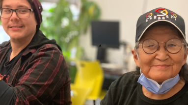 Two Indigenous participants sit at a table in CNIB Thunder Bay smiling. During a holiday program, they create decorations out of fresh cedar bows, dried orange slices, homemade gingerbread cookies, ribbons, bells, cinnamon sticks, and jute twine.