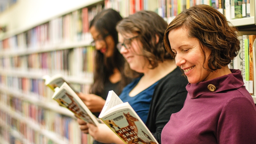 Three women standing in front of a row of bookshelves each reading a book.