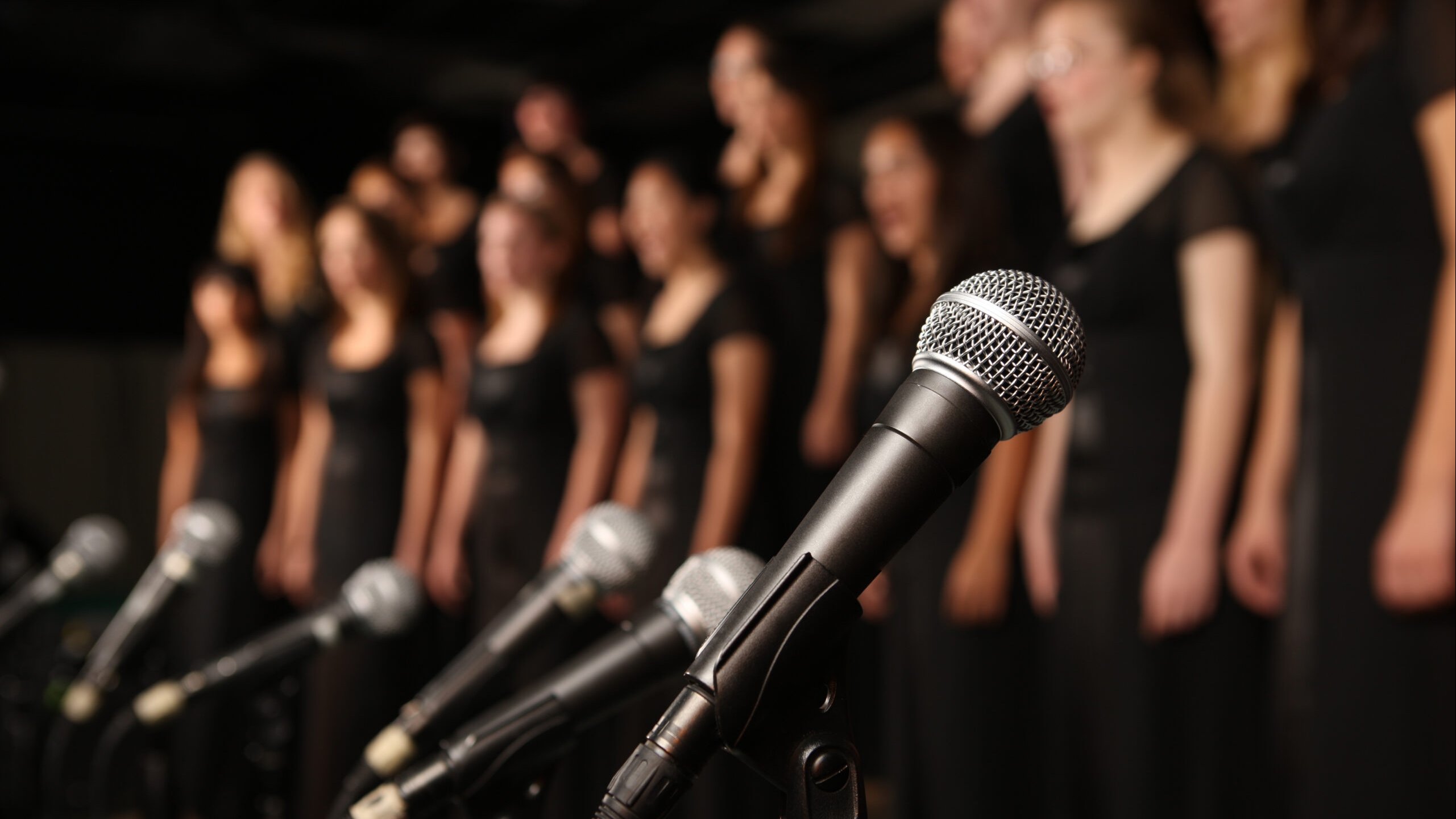 Photo of a number of microphones lined up with a group of women in black dresses in the background.