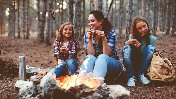 A woman and two girls sit outside in front of a fire eating treats.