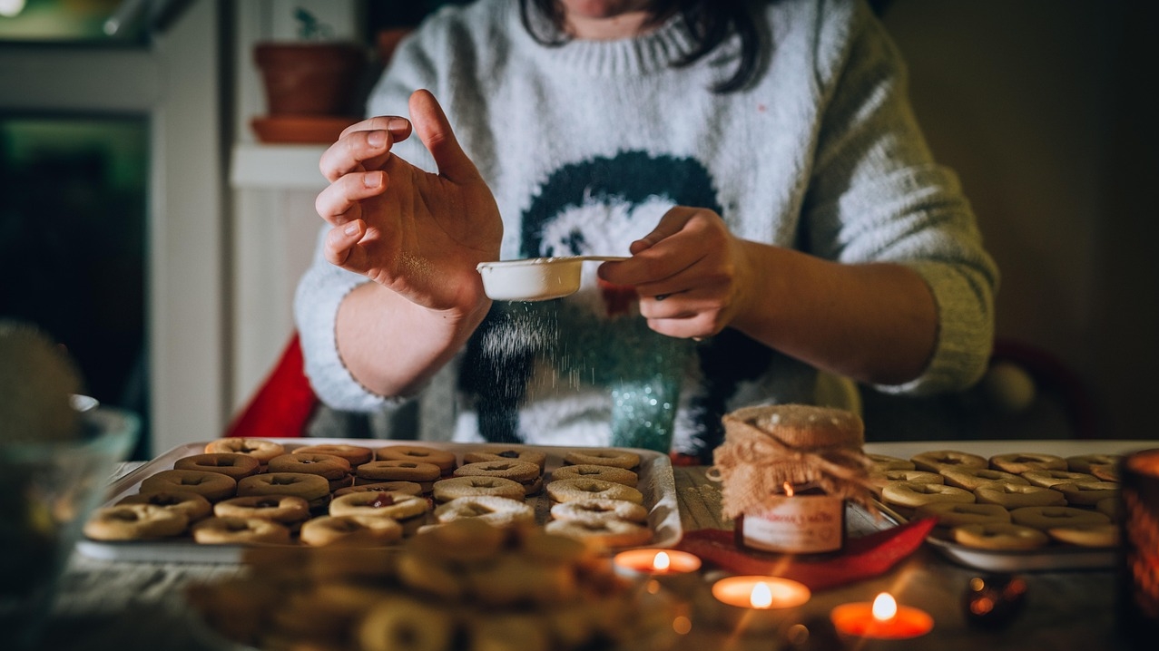 Woman sitting in front of a tray of baked goods. There are candles burning. She is sprinkling something on top.