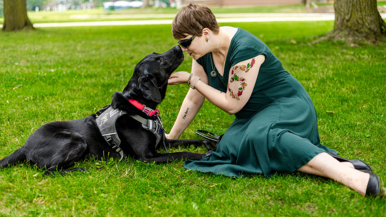 Denise and her guide dog Tara, a black Lab, sitting in the grass. Denise is gently holding Tara's face towards hers.