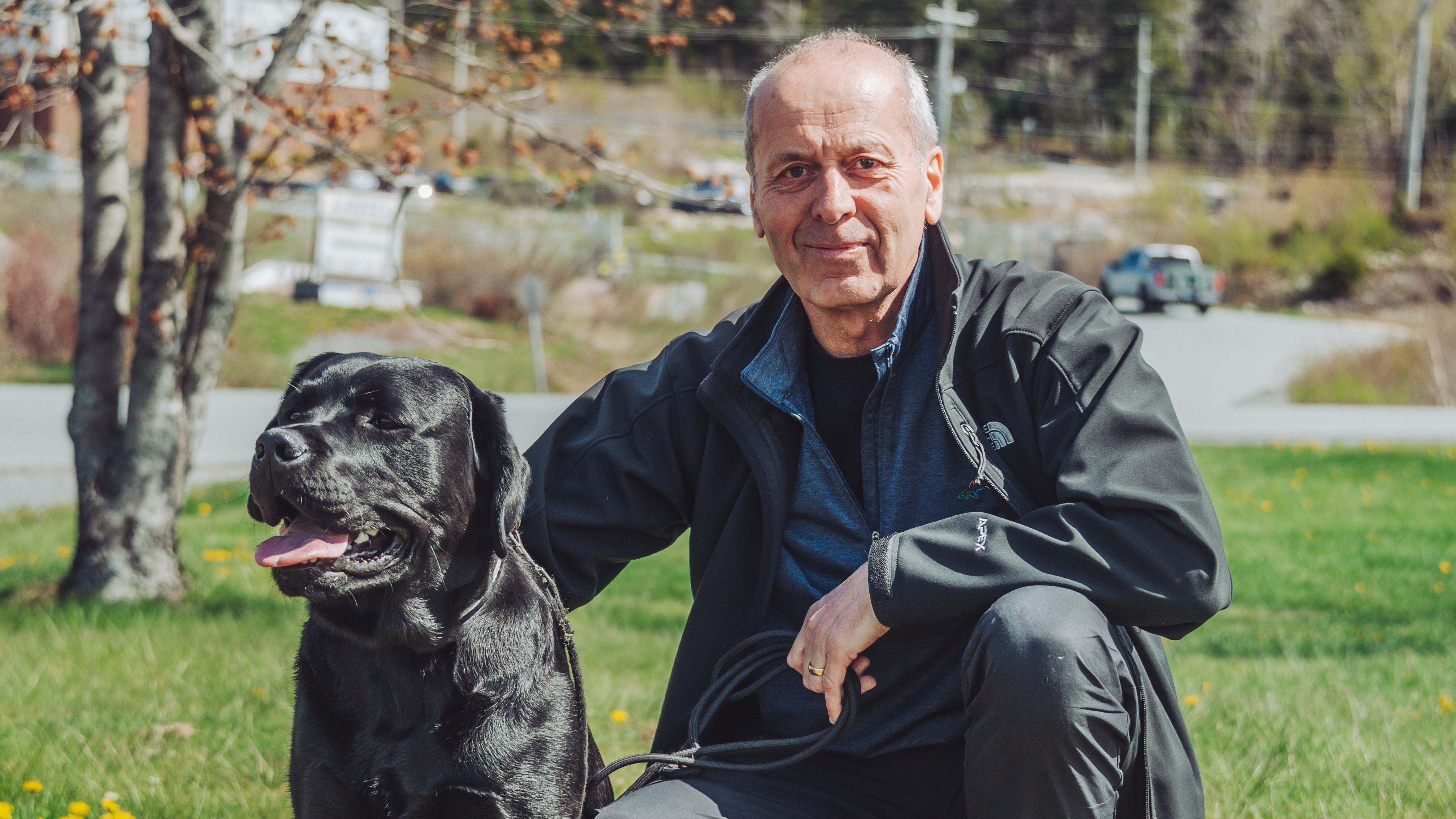 A man squats down beside a black Labrador retriever in a patch of grass. There is a tree with no leaves and a road behind them.