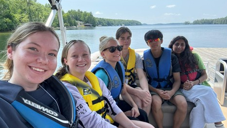 Young men and women are on a pontoon boat on the lake wearing colourful life jackets.