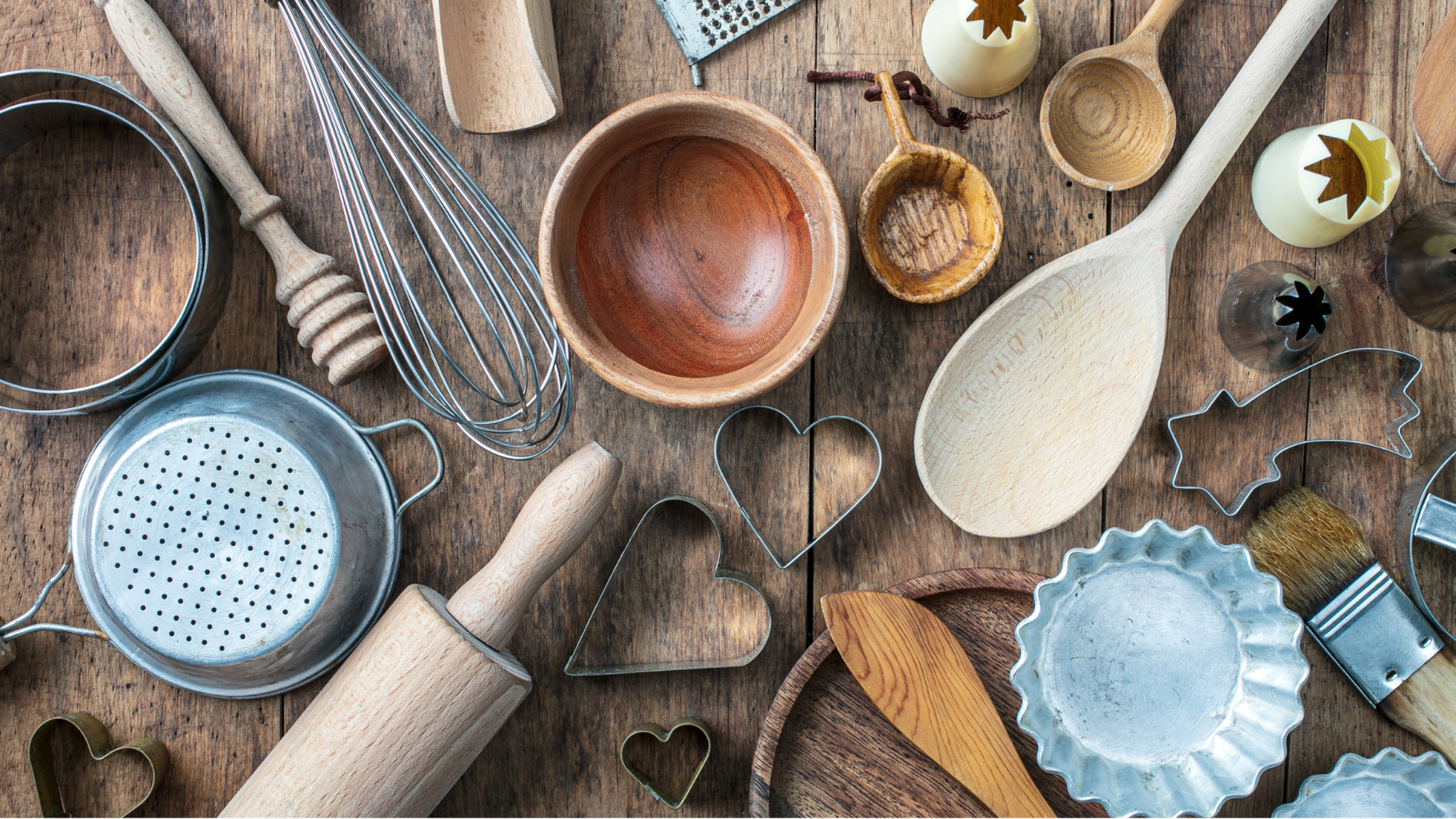 Bird's eye view of a variety of baking tools on a wooden table: spoons,, rolling pins, cookie cutters, etc.