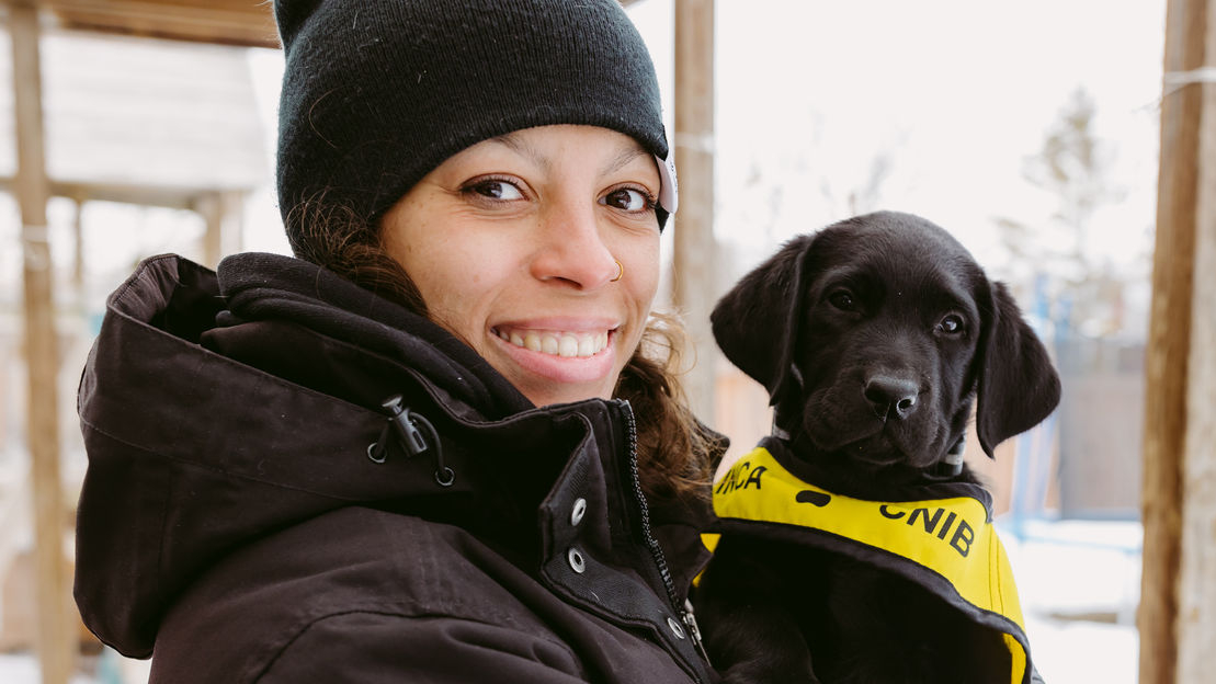 A smiling person in a winter jacket holds a black puppy wearing a CNIB Future Guide Dog vest.