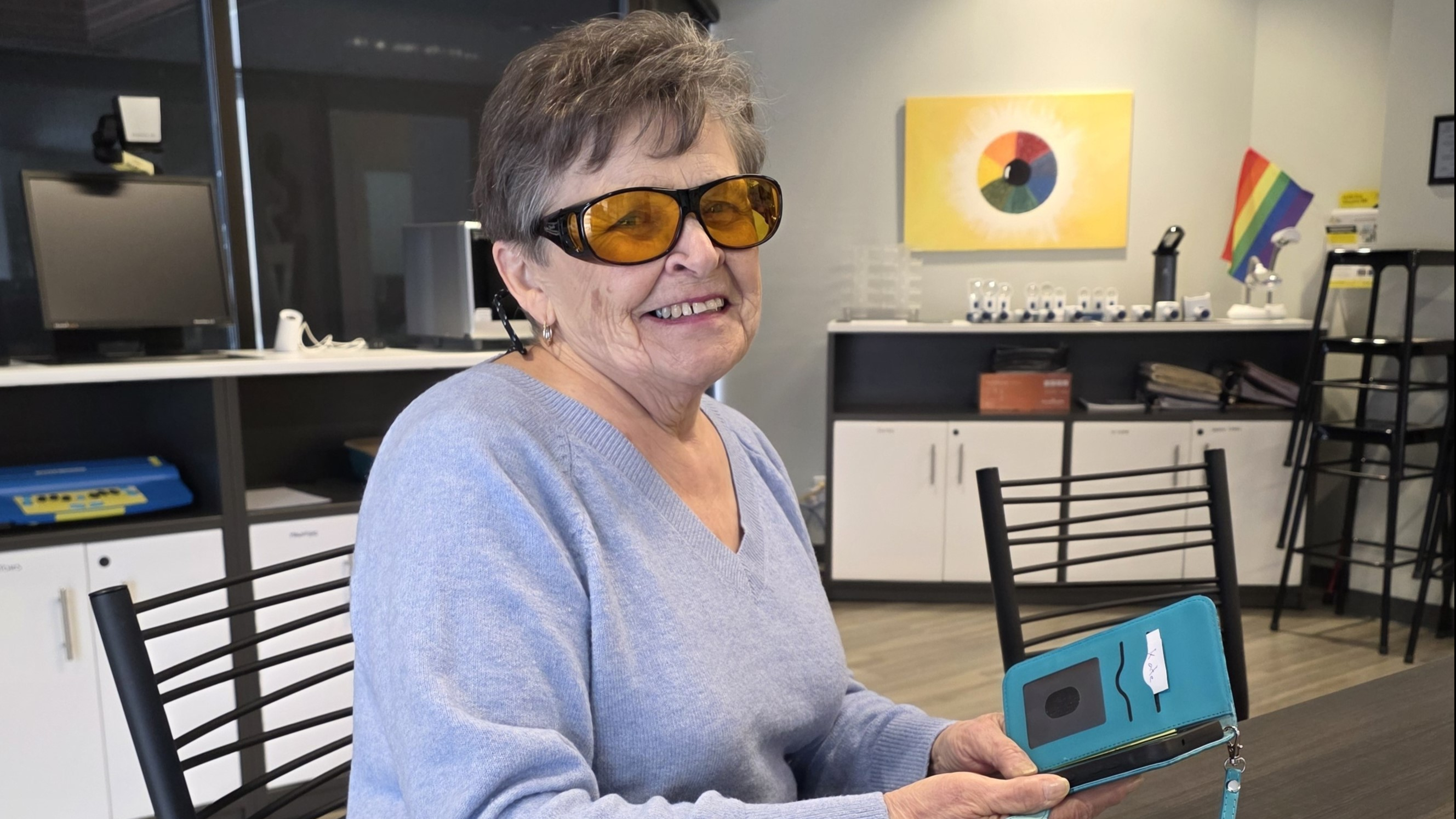 An older woman wearing amber-tinted glasses holding an accessible smartphone in a teal case.